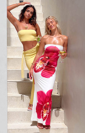 Two girls posing against a staircase in the sun. Brunette girl is wearing a yellow maxi dress and starfish necklace. Blonde girl is wearing a white maxi dress with a floral print.