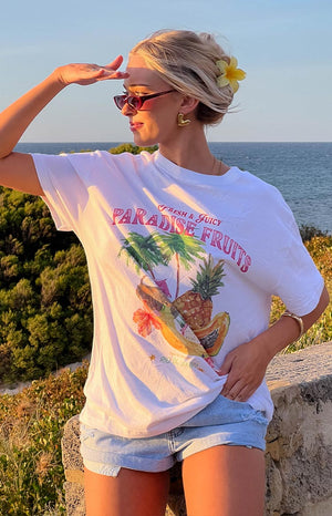 a blonde girl sits outside wearing a white printed t-shirt, sunglasses and jean shorts with a yellow flower clip in her hair