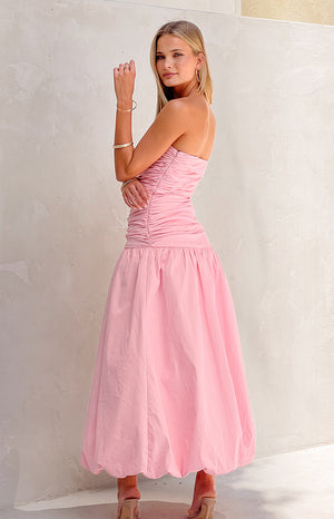 Girl wearing pink maxi dress with gold jewellery in front of white wall. 