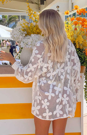 blonde girl wearing a white floral mesh long-sleeve top over a yellow bikini with her back turned