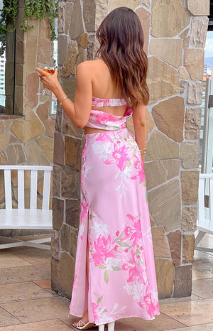 brunette model wearing a matching pink floral print maxi skirt and strapless top with cowl back, white heels and a gold bracelet holding a drink 