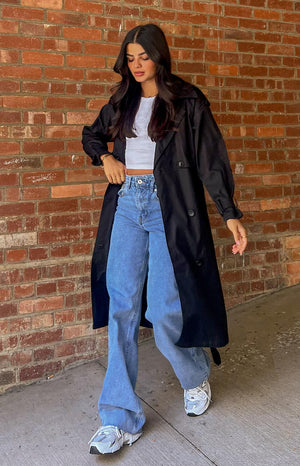 Brunette model is posed walking away from the camera, wearing a long, black trench coat. The trench coat features large black buttons on the centre and a fold over collar around the neck. 