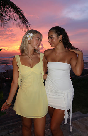 Two girls posing together in front of a sunset. GIrl is wearing a yellow mini dress with a white flower hair accessory. and girl is wearing a white strapless mini dress.