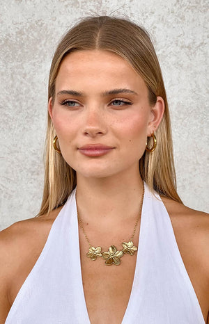 blonde model wearing a gold necklace with three gold flower pendants, gold hoop earrings and a white top