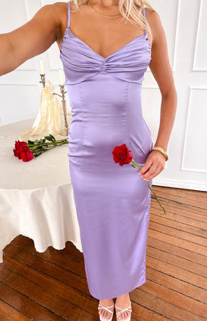 blonde girl wearing a purple maxi dress, gold bracelet and white heels holding a rose with a bouquette of roses on the table behind her