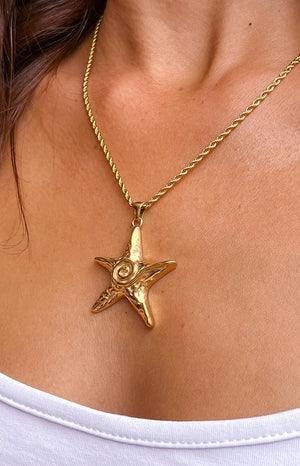 Close up image of a model with brunette hair wearing gold starfish necklace. The starfish pendant is attached to a gold rope chain. 