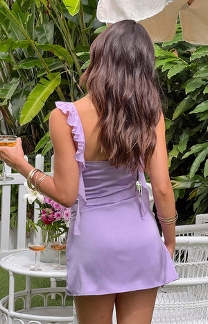 Girl facing away wearing purple mini dress in front of white table and garden.