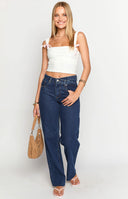 Model is wearing dark blue wash denim jeans with a straight leg fit and asymmetrical waist. Model is wearing brown sandals, a white top and is holding a brown leather bag.