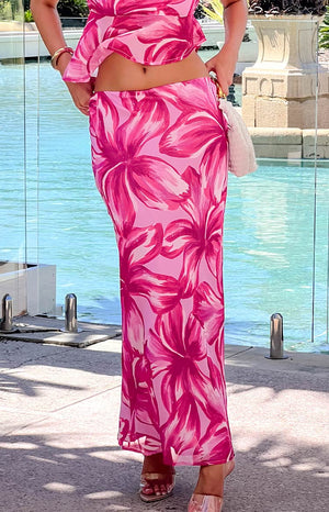 girl wearing a pink floral maxi skirt and top, holding a white purse beside a pool