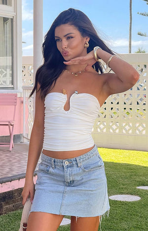 Brunette model is wearing white strapless top with a metal bar and orange stone detailing. Model is wearing a denim mini skirt and gold jewellery.