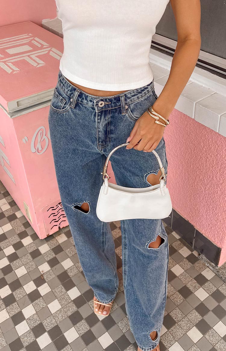 girl wears a white top, gold bracelets and blue denim jeans with love-heart cut outs holding a white purse