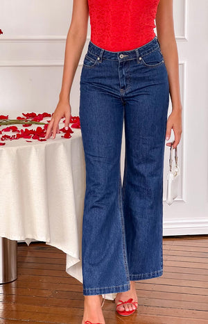 a girl wears dark blue denim jeans with a red top and red heels holding a white purse beside a table of rose petals