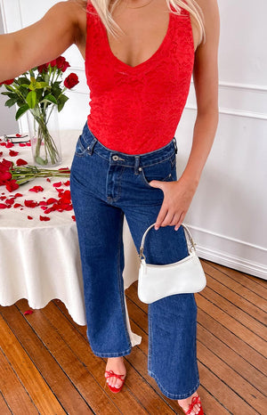 a blonde girl wears a red lace body suit, blue denim jeans, red heels holding a white purse beside a table of red roses