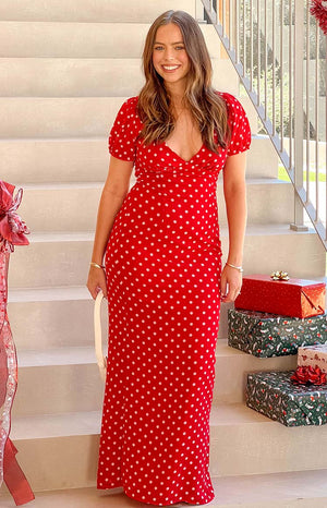 Brunette model wearing a red and white polka dot maxi dress with gold bracelets and holding a white hand bag.