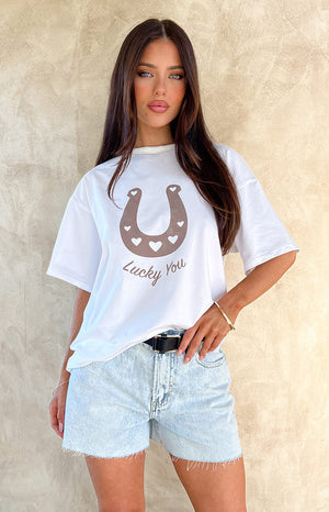 Brunette girl wearing a Lucky You White Western Tee and denim shorts standing in front of a wall.