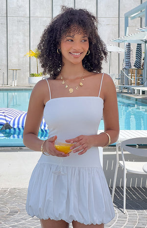 Girl wearing white mini dress and gold jewellery in front of pool area holding cocktail glass. 