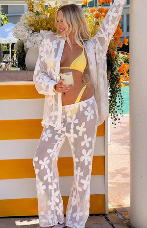 blonde girl wears a yellow bikini with a white floral top and pant set over top while holding a drink