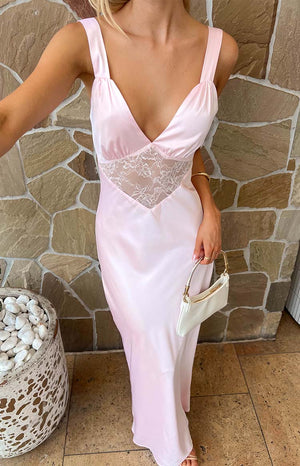 blonde model wearing a pink maxi dress with lace detailing in front and a gold bracelet holding a white purse beside a plant with white rocks