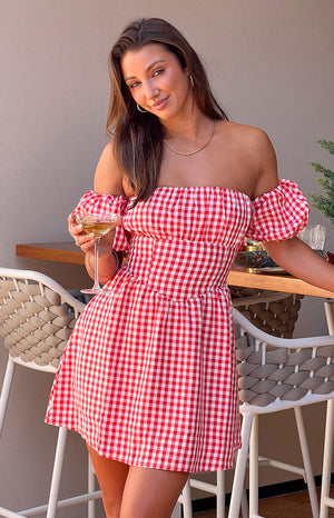 Brunette girl wearing red checkered mini dress holding wine glass in front of table.