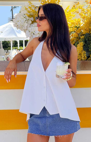 a brunette model wears a white button up top, sunglasses and denim mini skirt holding a drink