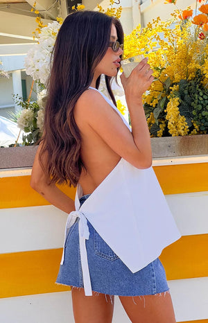 a brunette girl wears a white top with a low back, sunglasses and denim mini skirt holding a drink