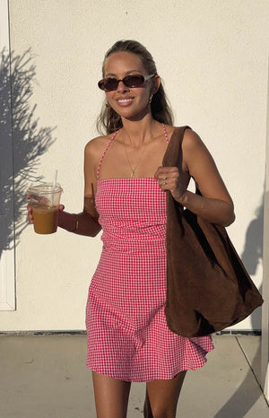 model is wearing a red gingham mini dress with a gathered bodice and brown bag 