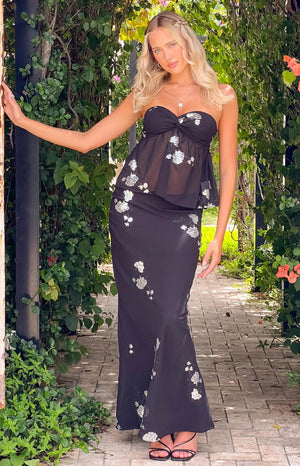 blonde girl wearing a black floral crop top and maxi skirt set and black heels posing with her arm raised