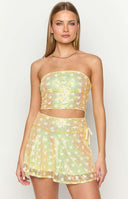 Girl in yellow sequin mini skirt and matching yellow top standing in front of pastel curtains.