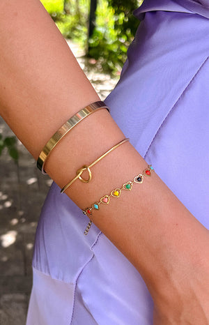 model wearing a purple satin dress with three gold bracelets, one thick bangle, one knotted cuff bracelet and one chain bracelet with rainbow heart charms