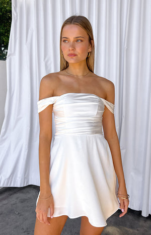 Girl wearing white off the shoulder mini dress in front of white curtains.