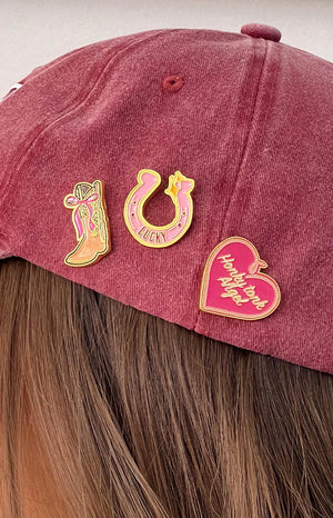 brunette girl wearing a red baseball cap with three western style pins - a cowboy boot with a bow, a pink lucky horseshoe and a pink heart with graphic text