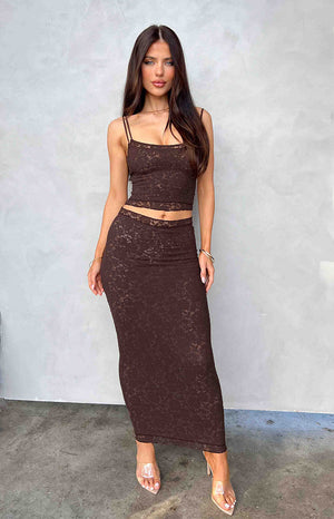 Brunette girl wearing a brown lace midi skirt and matching top standing in front of a wall.