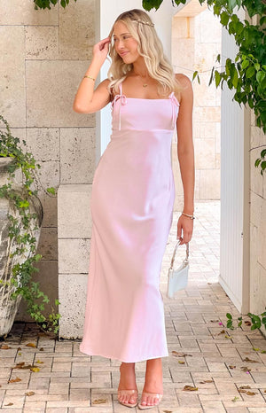 blonde girl wearing a soft pink maxi dress with bows on the straps, gold jewellery, nude heels holding a white purse