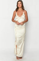 blonde model wearing a cream satin maxi dress with ruffle details, styled with a gold S initial necklace and white heels