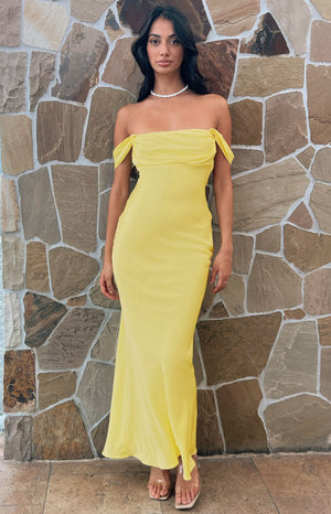 Model with black hair wears off the shoulder yellow maxi dress with nude heels and white beaded necklace.